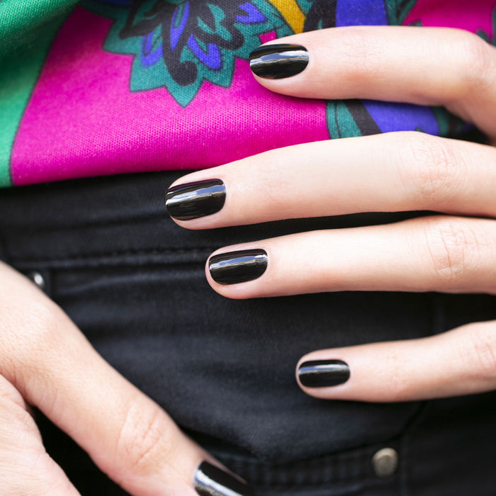 A close-up of a hand with Manucurist’s Licorice polish from the Green™ range, resting on black jeans against a vibrant pink and green patterned background.