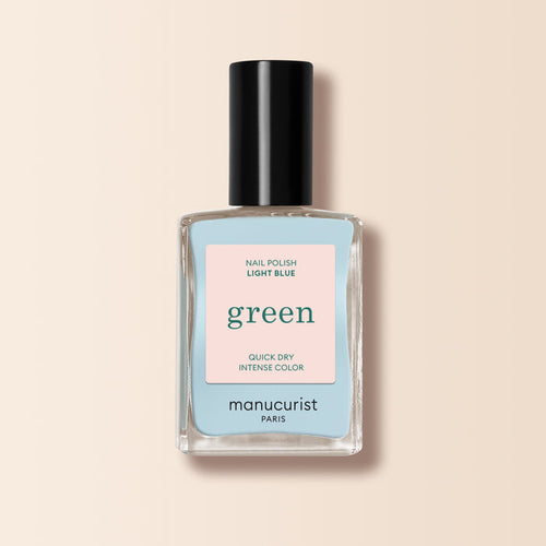A bottle of Manucurist Light Blue nail polish from the Green™ range, with a black cap and “green quick dry intense color” label, is shown against a beige background.