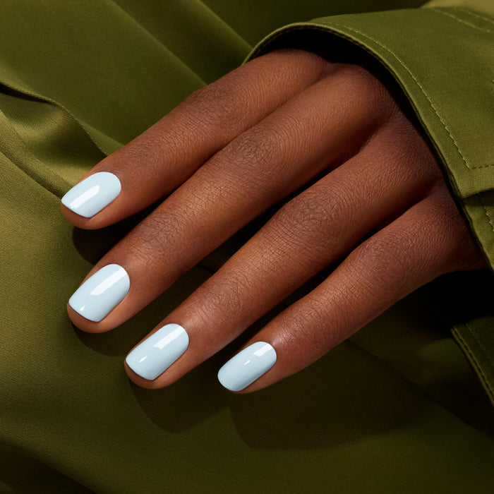 A hand with medium brown skin and neatly manicured nails painted in Manucurist Light Blue nail polish rests on olive green fabric.