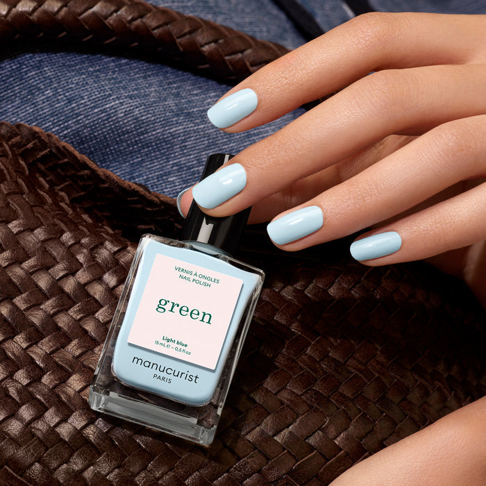 A hand with pastel light blue Manucurist nail polish holds a woven brown bag. A bottle of Manucurist Light Blue rests on the bag, with denim fabric in the background.
