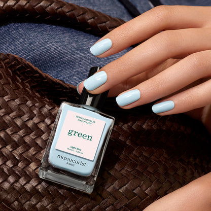 A hand with pastel light blue Manucurist nail polish holds a woven brown bag. A bottle of Manucurist Light Blue rests on the bag, with denim fabric in the background.