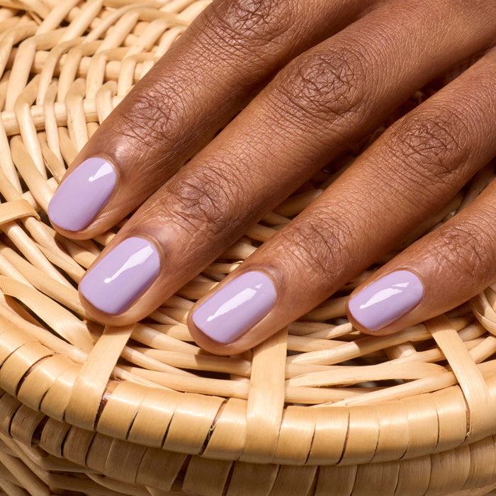 A hand with neatly manicured nails painted in Manucurist’s Lisa Lilas lilac nail polish rests gracefully on a woven wicker surface.