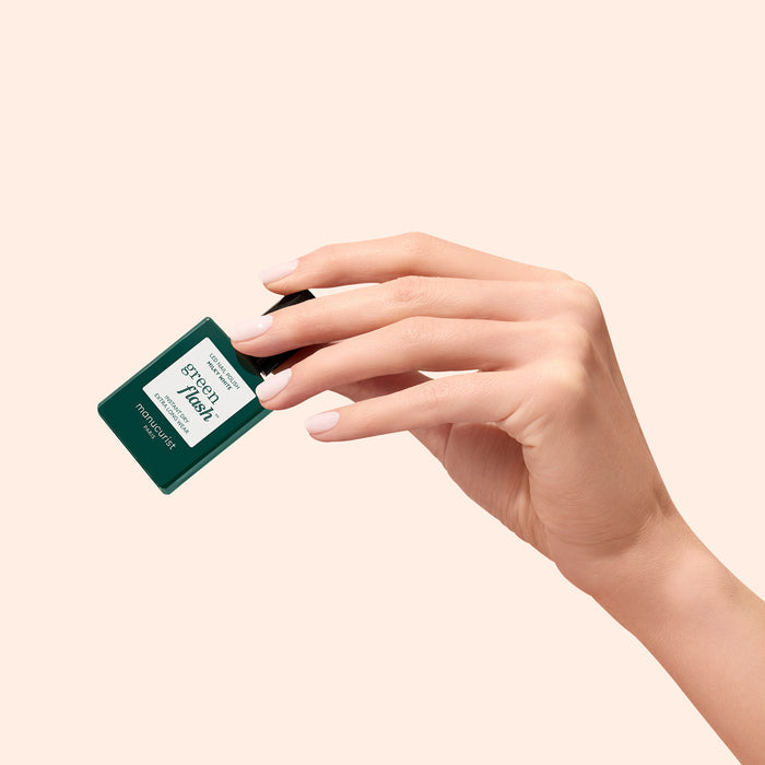 A hand with manicured nails holds a square, dark green Manucurist Green Flash gel polish bottle in Milky White against a light beige background. The label reads Step 1 and Manucurist.