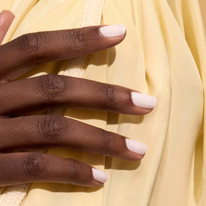 A close-up of a hand with medium-dark skin and neatly manicured nails painted in Milky White sheer gel polish by Manucurist, resting on soft, light yellow fabric.