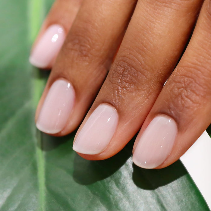 A close-up of a hand with short, neatly manicured nails painted in Manucurist’s Milky White sheer gel polish, resting on a green leaf.