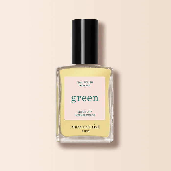 A bottle of Manucurist Mimosa pastel yellow nail polish from the Green™ range, featuring a black cap and label promoting its green quick-dry, intense color formula, is set against a soft beige background.