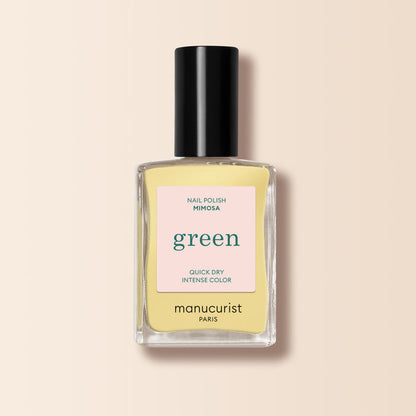 A bottle of Manucurist Mimosa pastel yellow nail polish from the Green™ range, featuring a black cap and label promoting its green quick-dry, intense color formula, is set against a soft beige background.
