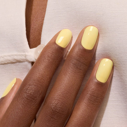 A close-up of a dark-skinned hand wearing Manucurist's Mimosa pastel yellow gel polish, resting on a white garment with button detail.