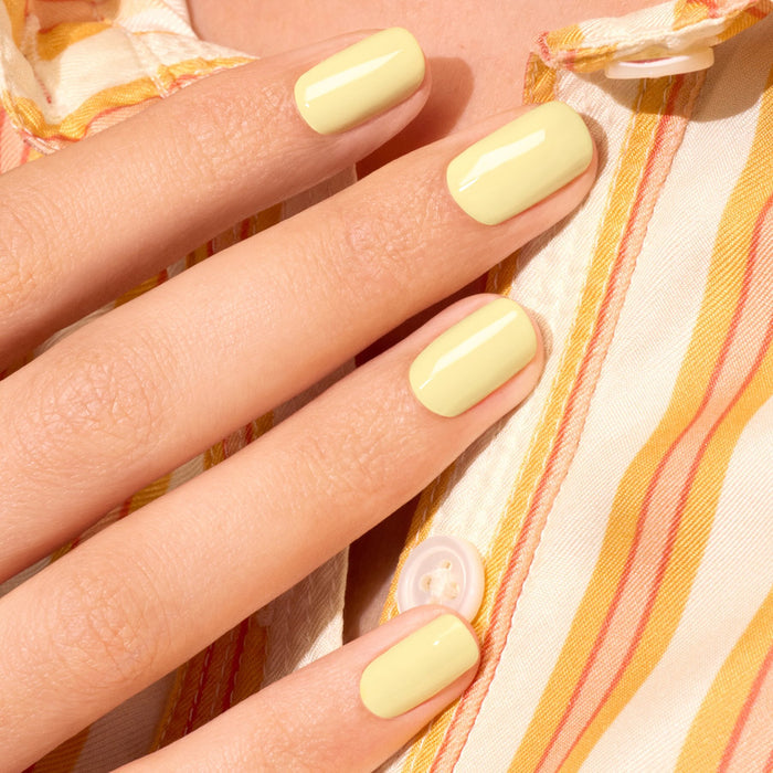 A hand with short, neatly filed nails painted in Manucurist’s Mimosa pastel yellow gel polish rests on a striped yellow, orange, and white shirt.