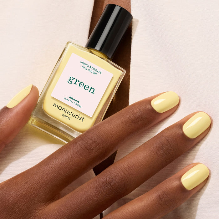 A hand with neatly manicured nails painted in pastel yellow holds a bottle of Manucurist Mimosa nail polish in a matching shade against a neutral background.