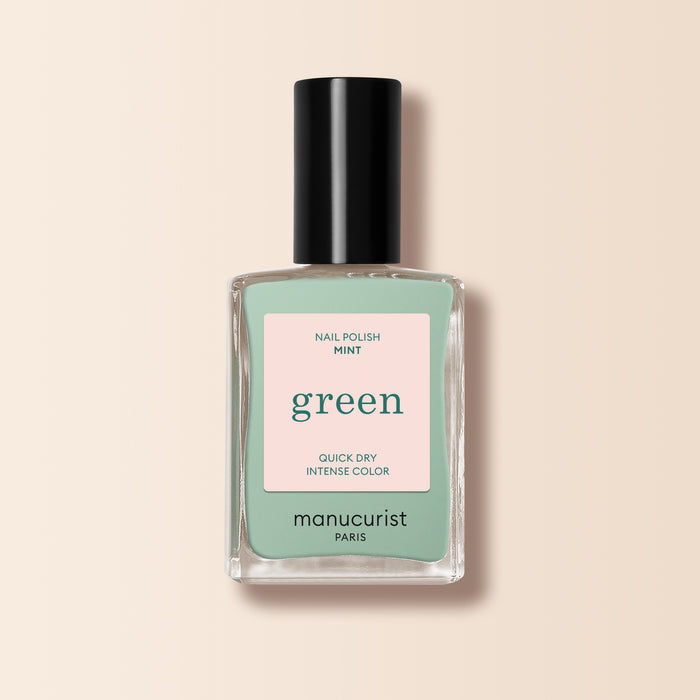 A bottle of Manucurist Mint nail polish from the Green™ line, pastel green with a black cap and a pink label reading "Nail Polish Mint, Quick Dry Intense Color," set against a pale beige background.