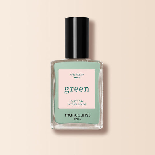 A bottle of Manucurist Mint nail polish from the Green™ line, pastel green with a black cap and a pink label reading "Nail Polish Mint, Quick Dry Intense Color," set against a pale beige background.