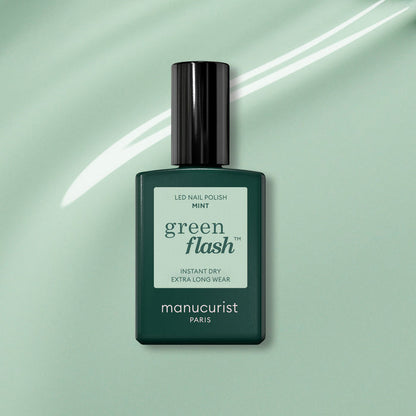 A bottle of Manucurist Mint Green Flash gel polish is displayed on a mint green background with a white swoosh. The dark green bottle features a black cap and a pastel green label.