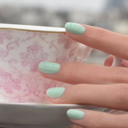 A hand with Manucurist's Mint pastel green gel polish holds a white teacup with a pink floral pattern, set against a blurred background.