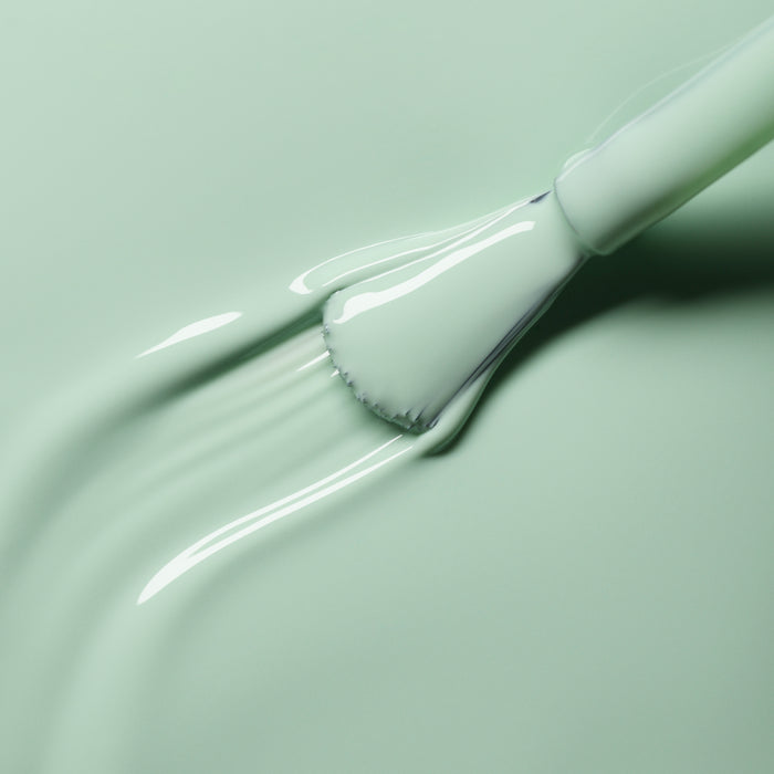 A close-up shows a brush spreading Manucurist's Mint nail polish over a smooth surface, with the bristles and handle partially visible against the creamy pastel green texture.