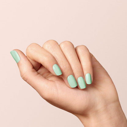 A light-skinned hand with neatly manicured nails is shown against a pale background, featuring pastel green "Mint" gel polish by Manucurist.