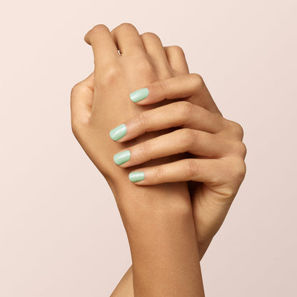 Two hands with light brown skin are gently clasped, displaying fingernails polished in Manucurist's Mint gel polish—a delicate pastel green—against a neutral beige background.