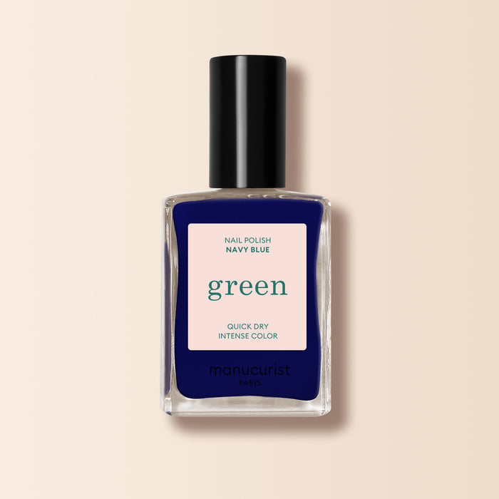 A bottle of Manucurist Navy Blue nail polish from the Green range with a black cap. The label says Nail Polish Navy Blue, Quick Dry Intense Color, and manucurist Paris. The background is soft beige.