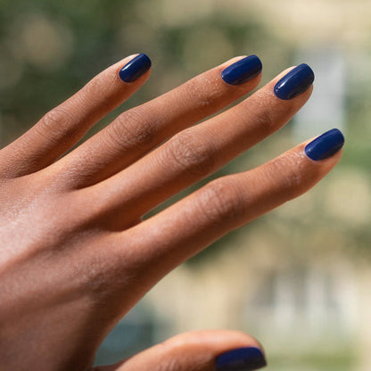 A hand with medium brown skin displays neatly manicured nails painted in Manucurist's Navy Blue polish. The fingers are spread, and the background features softly blurred greens and beige tones.