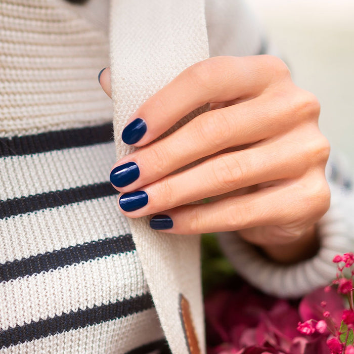 A hand with neatly manicured nails painted in Manucurist Navy Blue rests on a cream knit scarf. The person wears a white sweater with black stripes, and pink flowers can be seen in the background.