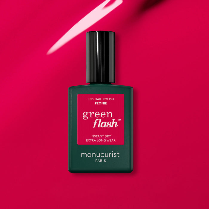 A bottle of Manucurist Péonie Green Flash gel polish rests on a glossy, bright pink surface reflecting light.