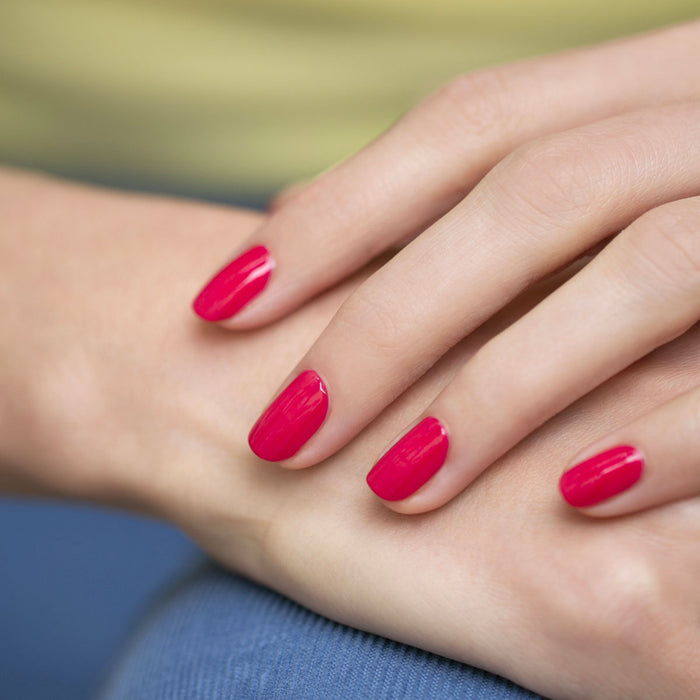 A close-up of a hand with neatly manicured nails painted in Manucurist's Péonie, a raspberry pink gel polish, gently resting on the skin against a softly blurred blue and yellow background.