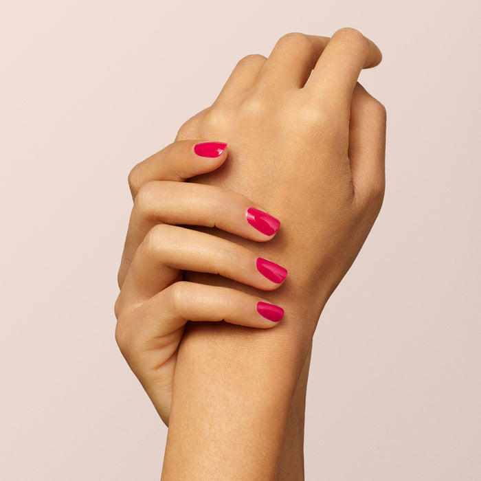 A close-up of two hands with neatly manicured nails painted in Manucurist Péonie, a vibrant raspberry pink gel polish, set against a soft, light beige background.