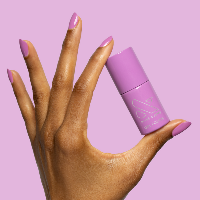 Hand with nails painted in bright floral purple shade holding Petal - Gel polish bottle on purple background