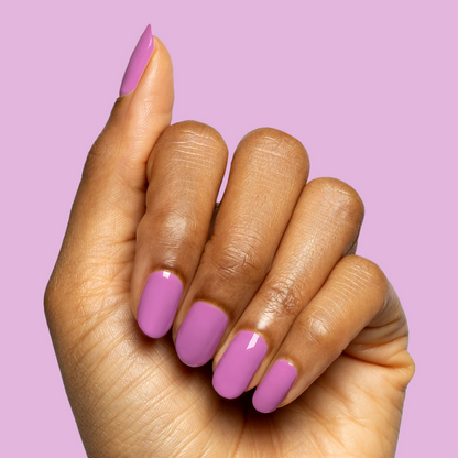 Hand with rounded nails painted in bright floral purple shade using Petal - Gel on soft pink-purple background
