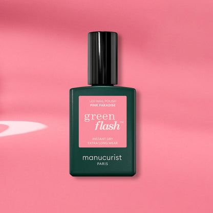 A Manucurist Pink Paradise Green Flash gel polish bottle, with a dark green base and black cap, is displayed on a pink background with soft shadows.