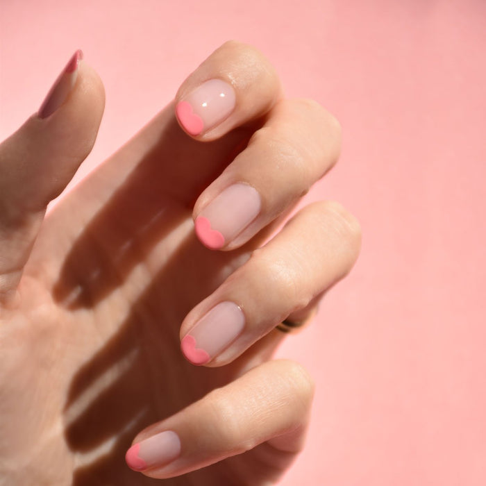A hand with manicured nails shows a minimalist look: each nail is painted clear with light pink heart-shaped tips using Manucurist’s Pink Paradise gel polish, set against a soft pink background.