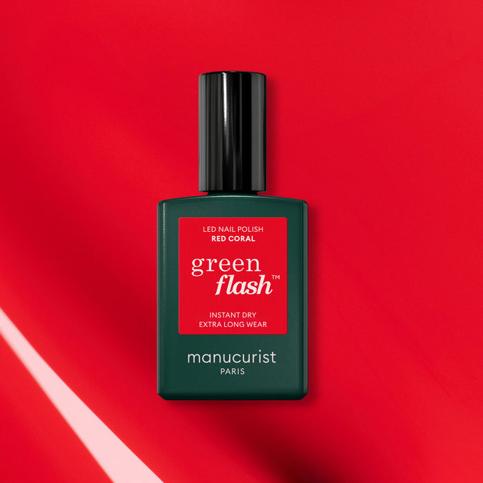 A bottle of Manucurist Red Coral Green Flash gel polish rests on a vibrant red background with diagonal light reflections, its dark green base and red label highlighting a festive red nail polish look.