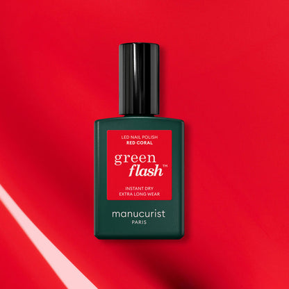 A bottle of Manucurist Red Coral Green Flash gel polish rests on a vibrant red background with diagonal light reflections, its dark green base and red label highlighting a festive red nail polish look.