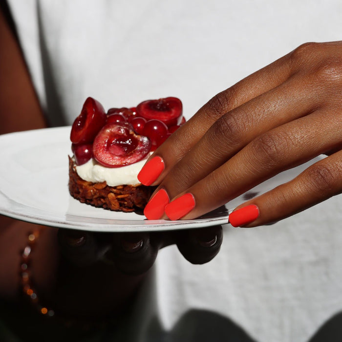 A hand with Manucurist's Red Coral nail polish holds a white plate with a dessert topped with whipped cream and fresh cherries. The person is wearing a white shirt.
