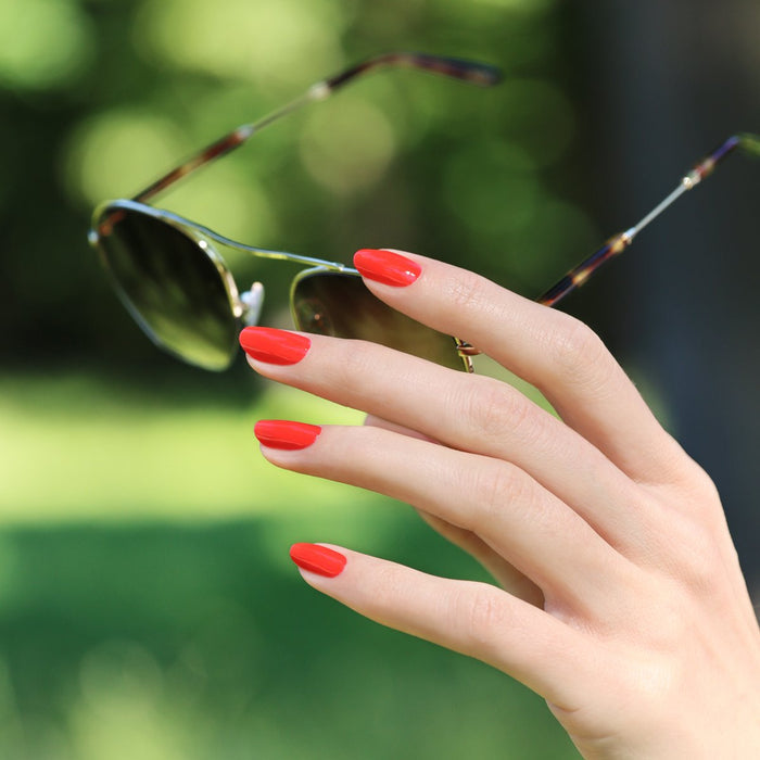 A hand with neatly manicured nails painted in Manucurist's Red Coral polish holds sunglasses outdoors against a blurred green background.