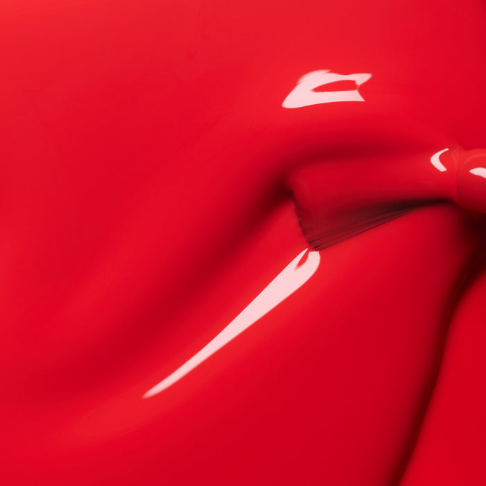 Close-up of Manucurist Red Coral gel polish: a glossy, smooth coral-red surface with a shiny highlight and visible brush stroke, evoking the look of freshly applied nail polish.