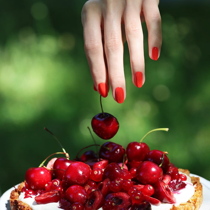 A hand with Manucurist's Red Coral nail polish holds a cherry above a tart topped with cherries and red berries, set against a blurred green outdoor background.