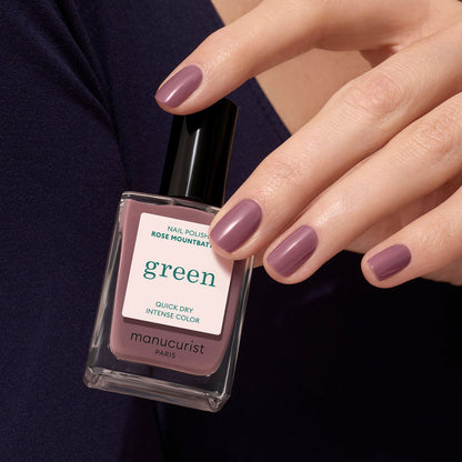 A hand with short, neatly manicured nails painted in Manucurist’s Rose Mountbatten holds the matching bottle from their Green™ range.