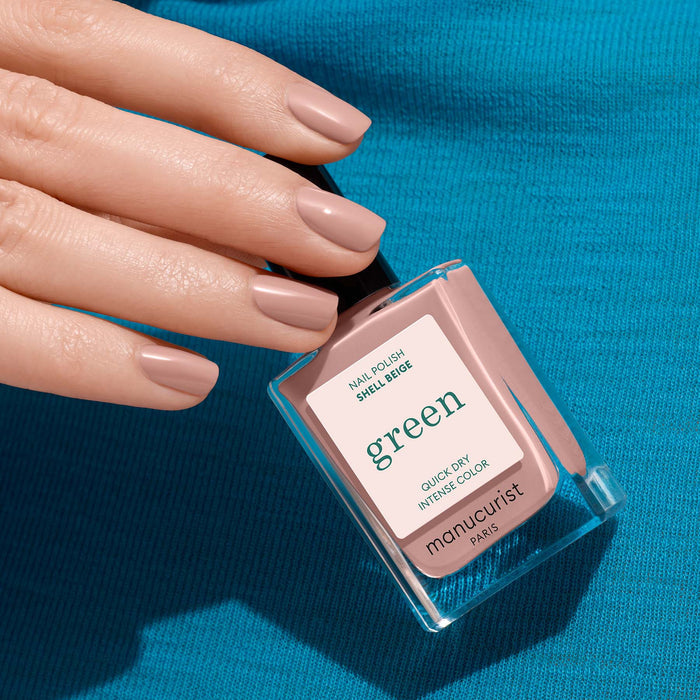 A hand with neatly manicured nails in a delicate pinkish beige shade holds a square bottle of Manucurist Shell Beige nail polish against a bright blue fabric background.