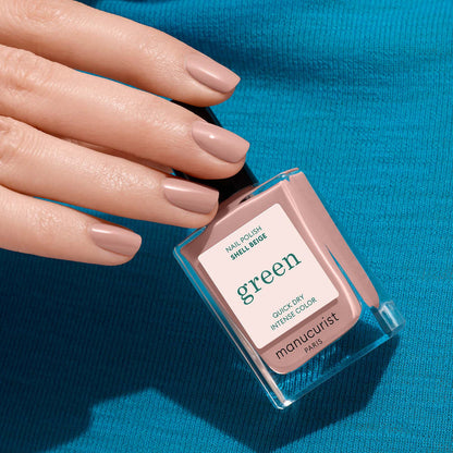 A hand with neatly manicured nails in a delicate pinkish beige shade holds a square bottle of Manucurist Shell Beige nail polish against a bright blue fabric background.