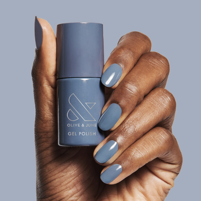 Hand holding Olive & June Slate - Gel polish bottle with matching midtone slate blue manicure on natural skin tone