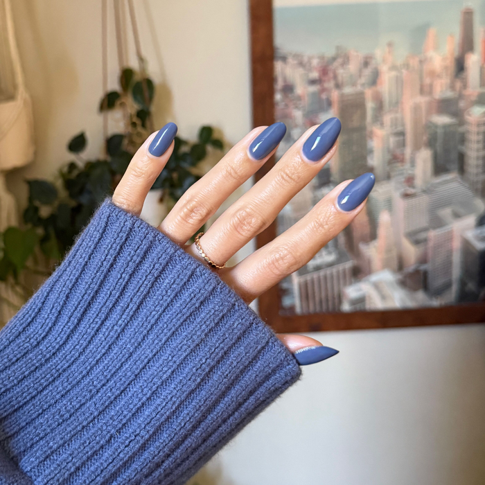 Hand with nails painted in Slate - Gel, a smooth midtone slate blue gel polish, wearing a matching blue sweater sleeve