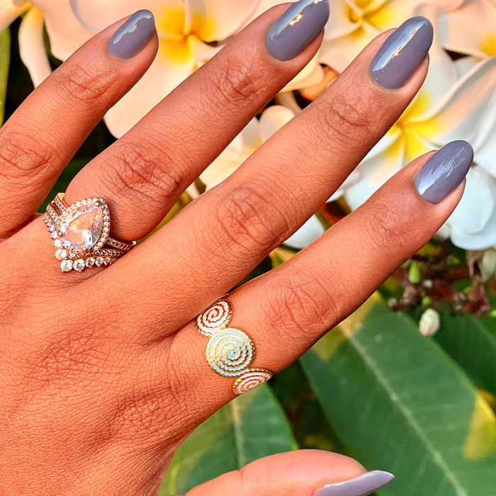 Hand with nails painted in Slate - Gel, a midtone slate blue gel polish, adorned with rings among tropical flowers and leaves