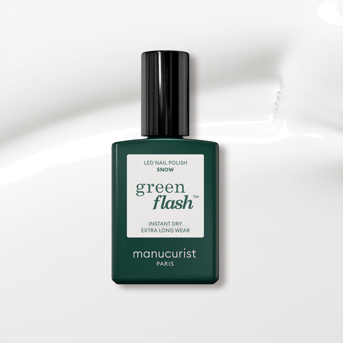 A dark green Manucurist bottle of Green Flash gel polish in the shade Snow sits on a glossy white surface. The label highlights instant dry and extra long wear, ideal for achieving a pure white nail polish look.