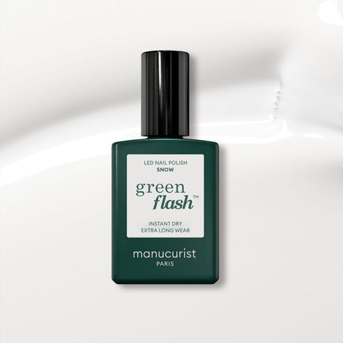 A dark green Manucurist bottle of Green Flash gel polish in the shade Snow sits on a glossy white surface. The label highlights instant dry and extra long wear, ideal for achieving a pure white nail polish look.