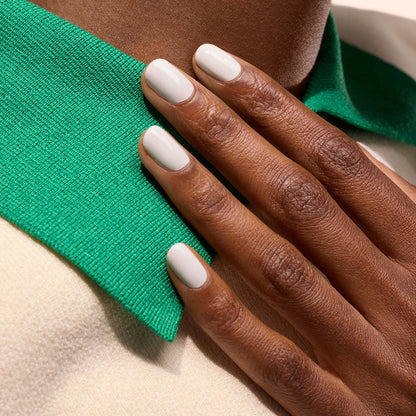A hand with neatly manicured, short nails coated in Manucurist's Snow pure white nail polish rests on a green collar of a cream-colored garment.