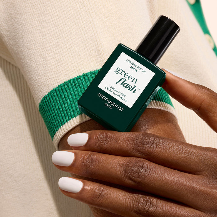 A hand with pure white "Snow" nail polish by Manucurist holds a dark green bottle of Manucurist Green Flash gel polish against a cream sweater with green trim.