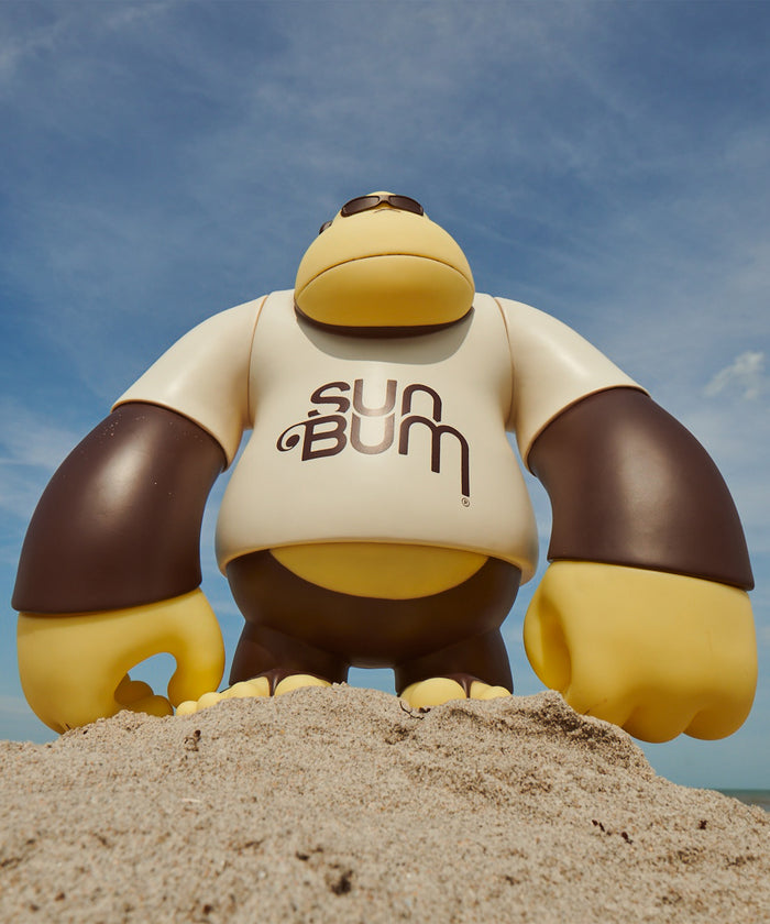 Giant Sun Bum mascot figure standing on sandy beach under blue sky - Brand Vibes Collection - Sun Bum