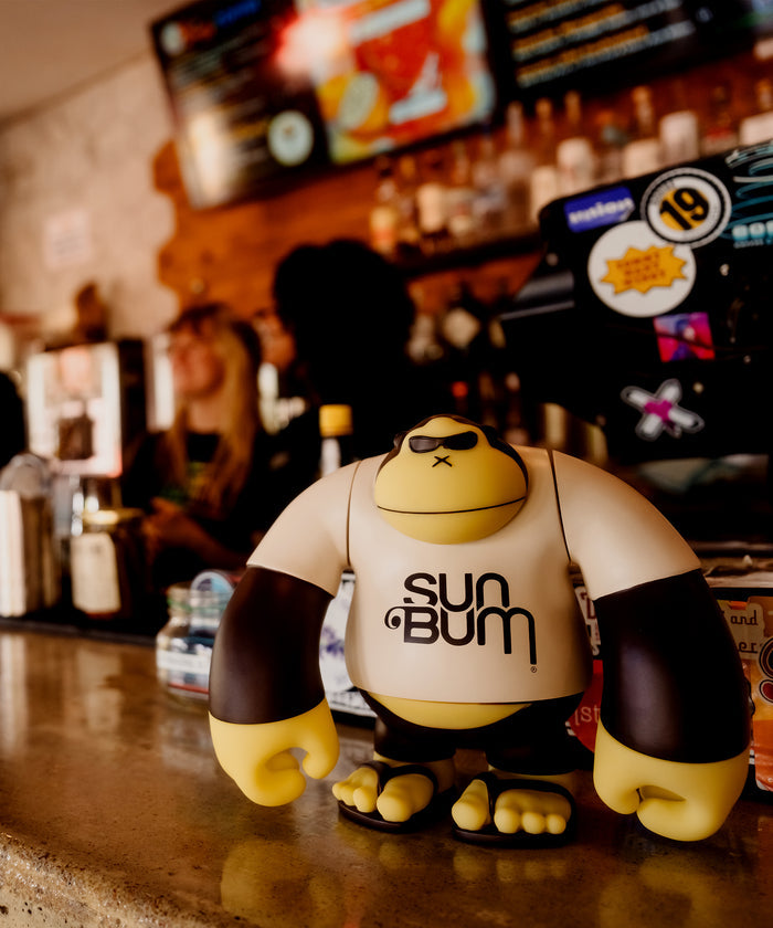 Sonny' 9" Vinyl Figure by Sun Bum wearing sunglasses and a branded shirt standing on a bar counter
