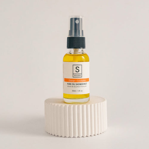 A small white spray bottle labeled SoulDeo Shower Mist with orange and eucalyptus, positioned on a textured white surface against a neutral background.